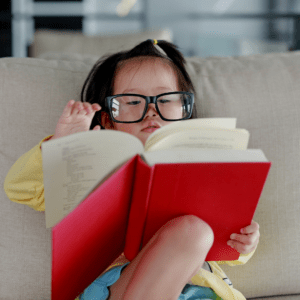 Asian girl sitting on a couch reading a book while wearing glasses